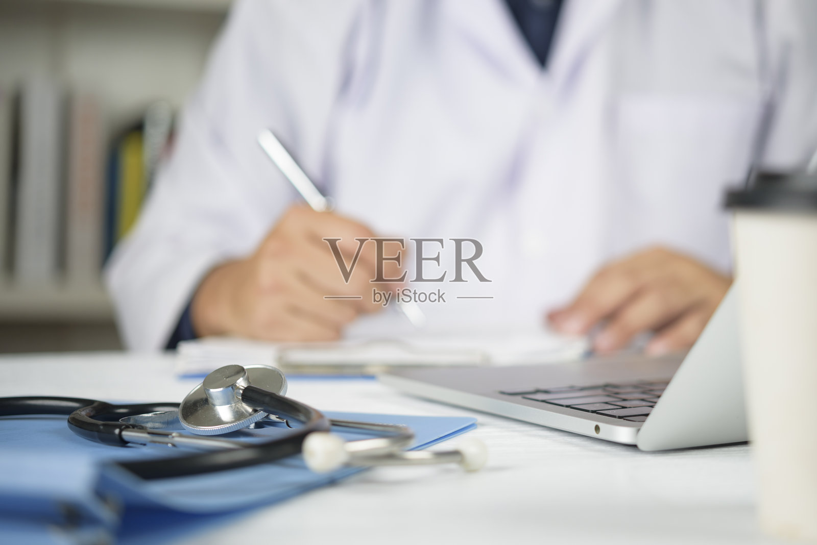 桌上的听诊器，背景是医生在写病人报告。医生在医院办公室工作，使用笔记本电脑进行研究和远程医疗。专业的医疗咨询概念。照片摄影图片