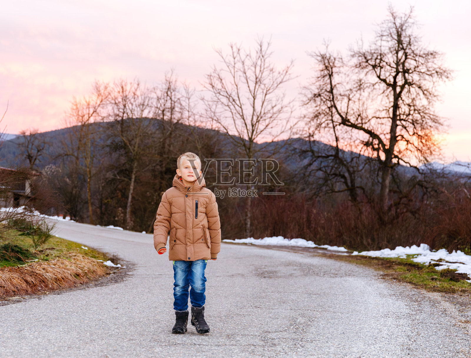 日落时分，身穿棕色夹克的白种男性儿童站在乡村的冬日公路上照片摄影图片