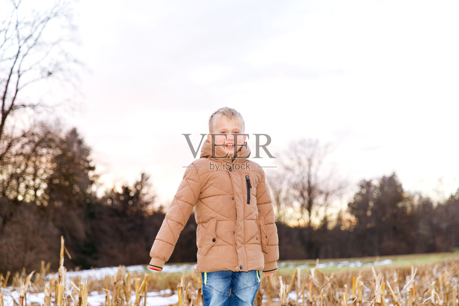 穿着棕色外套的金发小男孩在日落时分的冬日田野里站着照片摄影图片