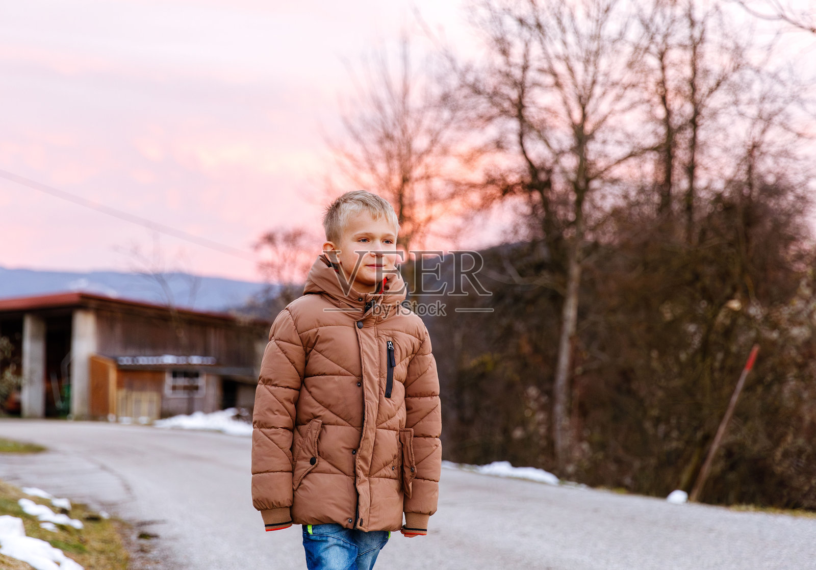 日落时分，一个穿着棕色夹克的白人小男孩走在乡村小路上照片摄影图片