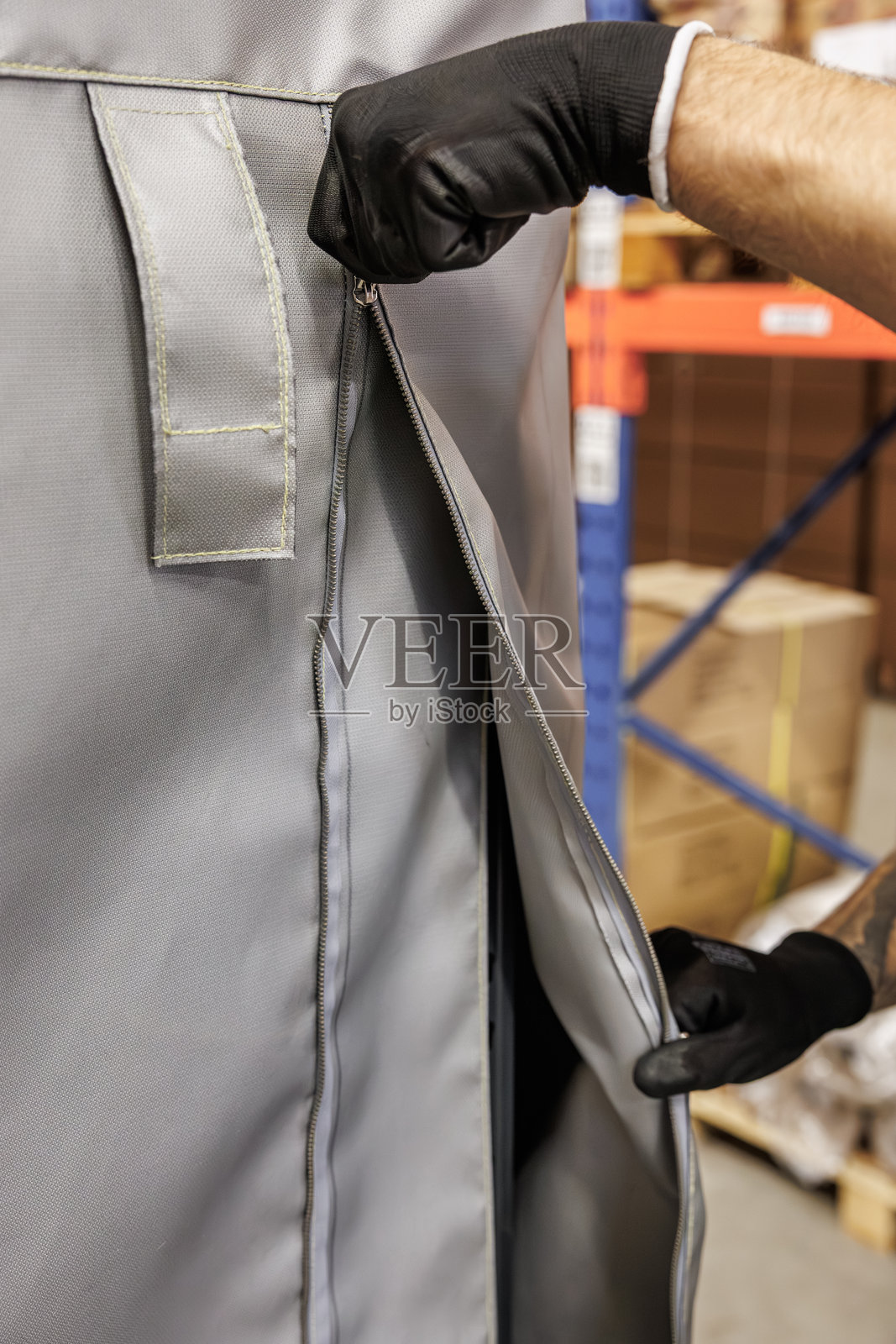 在仓库环境中，特写镜头展示了一只戴着黑色手套的手正在拉开厚实的灰色工业织物罩的拉链。背景中可以看到纸箱和货架。照片摄影图片