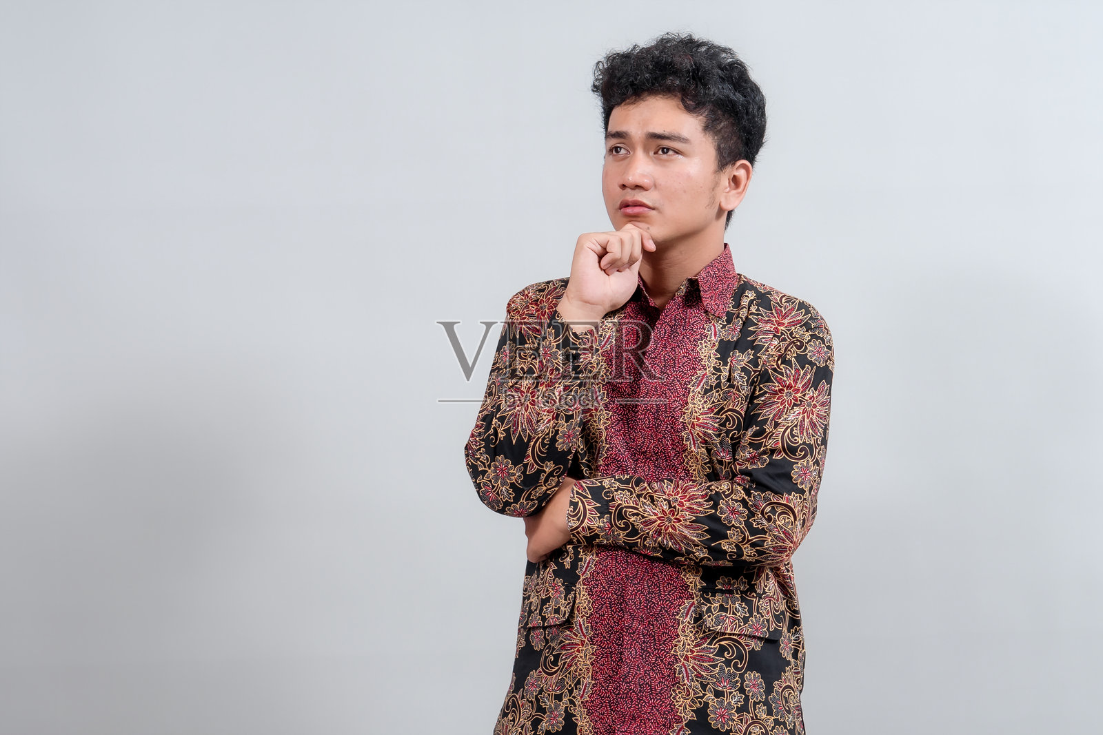 一位身穿传统蜡染衬衫的年轻男子若有所思地摆出姿势，手托着下巴，在纯色背景下流露出沉思的神情。照片摄影图片