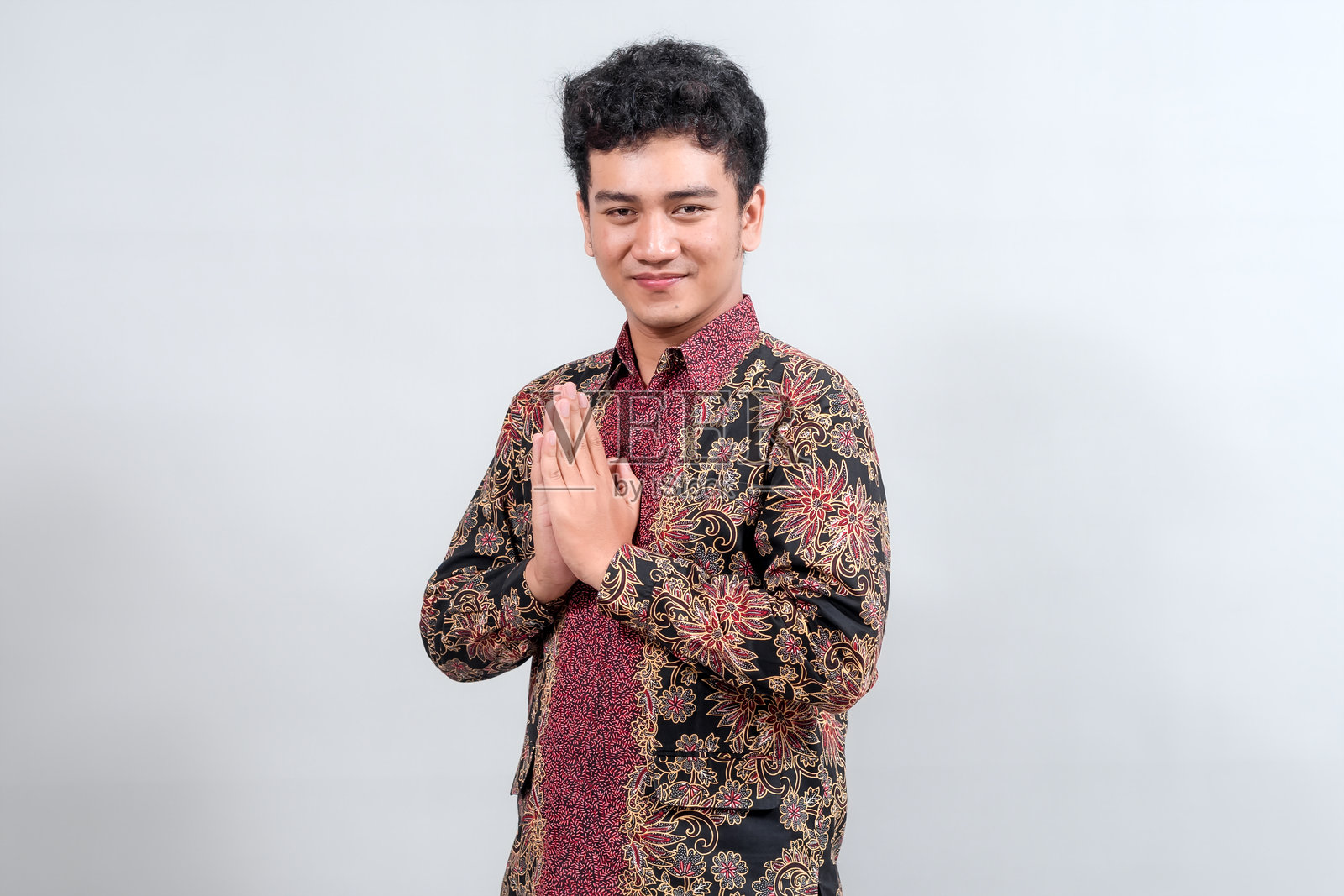一位身穿传统蜡染衬衫的年轻男子双手合十，做出祈祷姿势，面带温暖的微笑，背景中性，展现出文化尊重。照片摄影图片