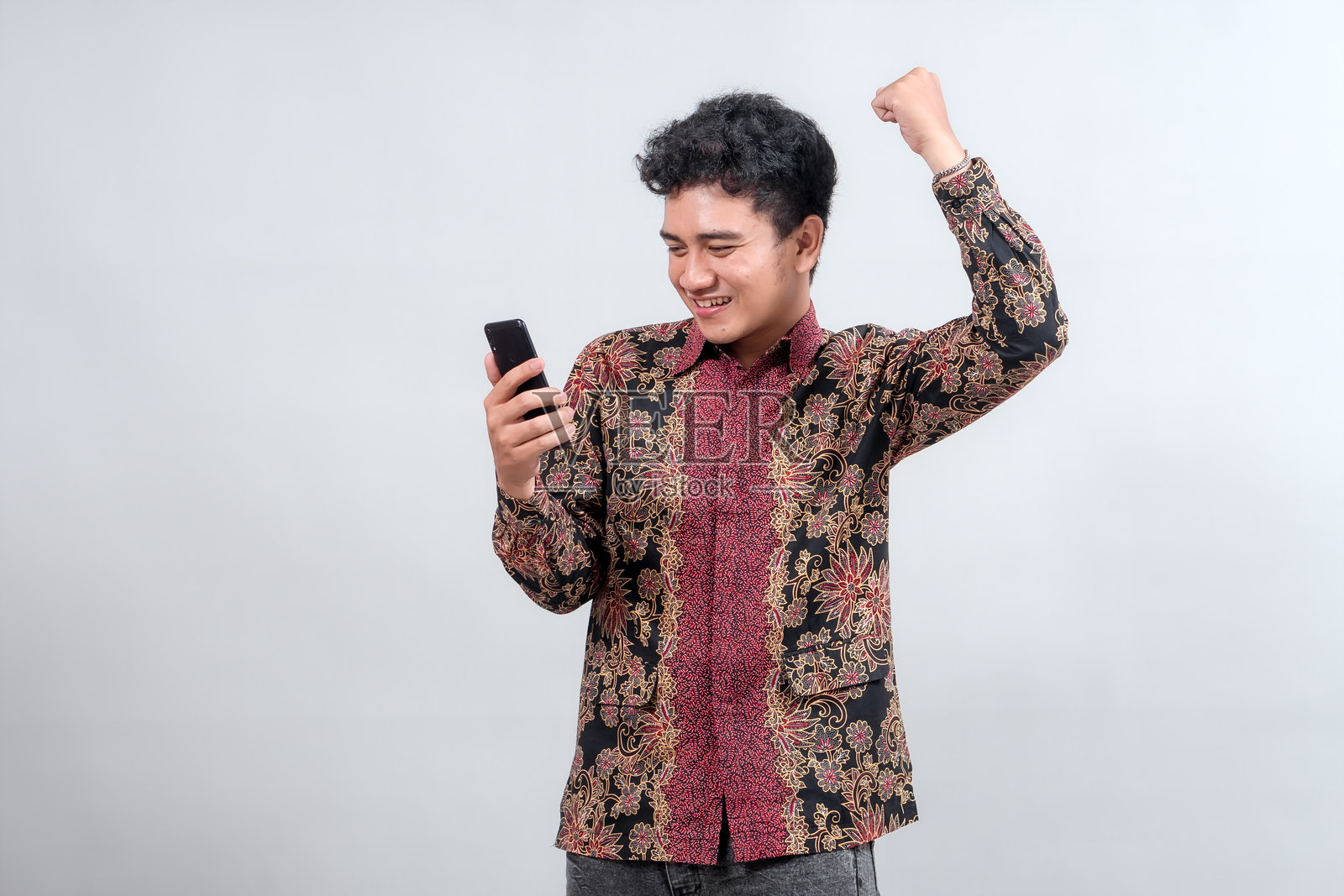一位穿着鲜艳蜡染衬衫的年轻男子，在现代数字背景下，喜悦地庆祝着，并兴奋地看着他的智能手机，表达着胜利的喜悦。照片摄影图片