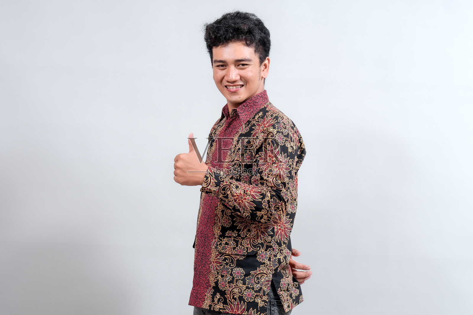 一位身穿色彩鲜艳的蜡染衬衫的年轻男子，自信地竖起大拇指并微笑，展现了印度尼西亚传统时尚与现代积极的融合。照片摄影图片