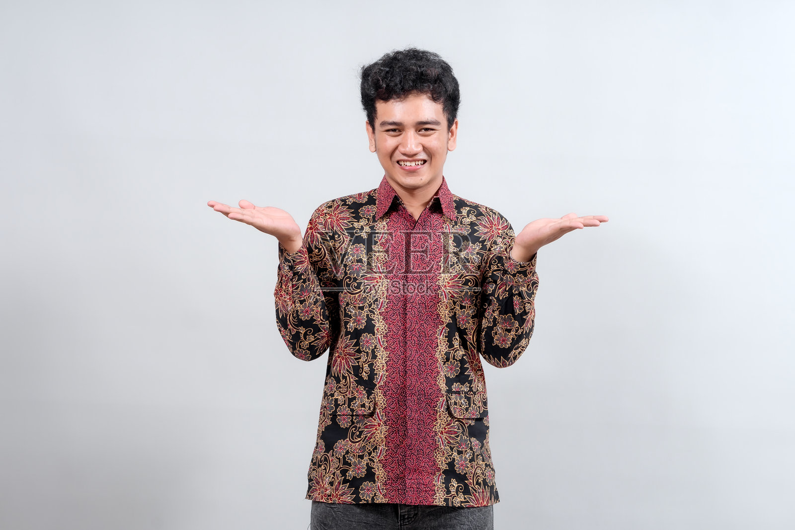 一位身穿色彩鲜艳的蜡染衬衫的快乐年轻男子，双手张开呈展示状微笑着，展现了印度尼西亚传统时尚与友好风度的融合。照片摄影图片