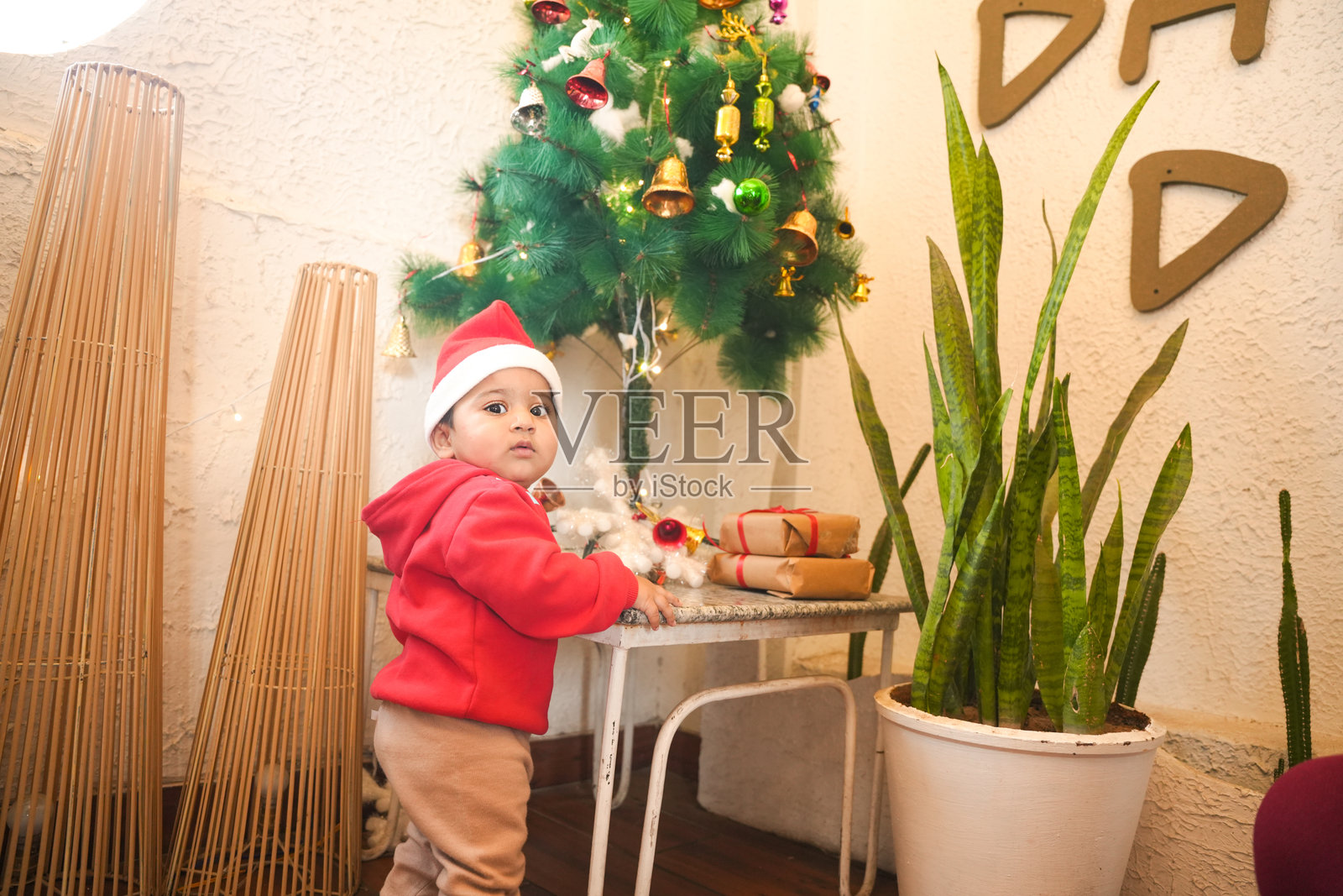 家里装饰着节日装饰的圣诞树，一个戴着圣诞帽的男婴站在旁边。照片摄影图片