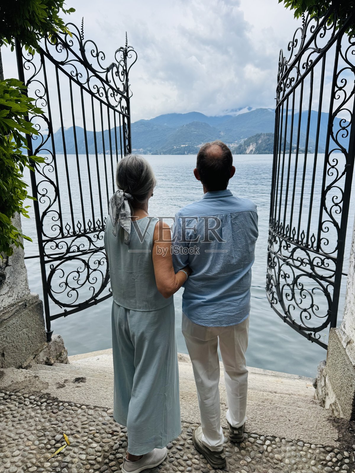 一对年长的夫妇并肩站立，透过意大利科莫湖边的大型敞开式铁门向外眺望。背影。照片摄影图片