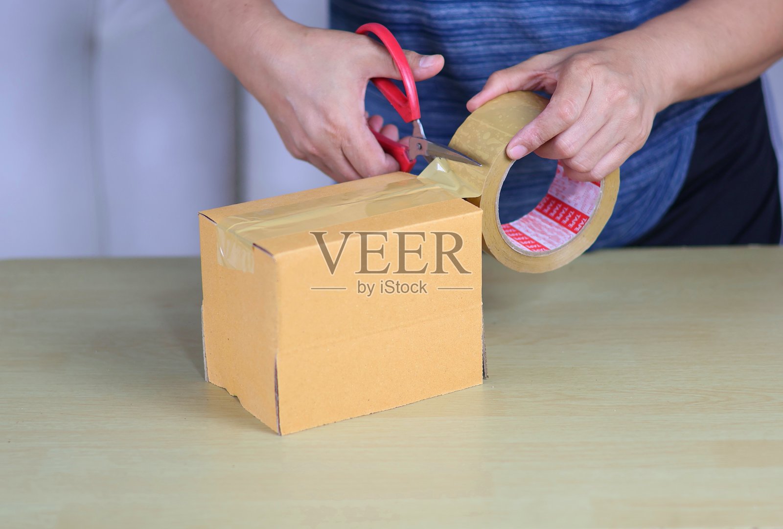 一个人用红色剪刀剪下透明胶带，封住一个棕色纸箱。焦点集中在手和纸箱上，背景为中性色。照片摄影图片