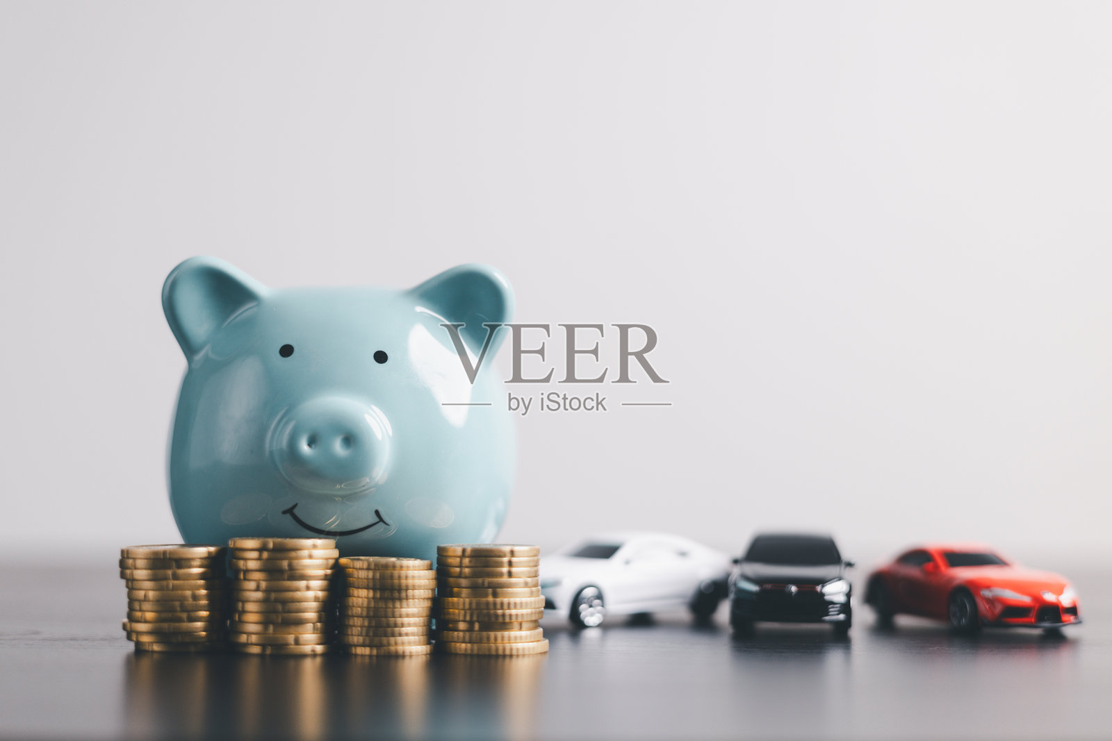 一个蓝色的储蓄罐，旁边堆叠着硬币，还有三个汽车模型，在干净简约的背景上，代表着储蓄、汽车贷款、金融、投资、财富管理、保险和金钱规划。照片摄影图片
