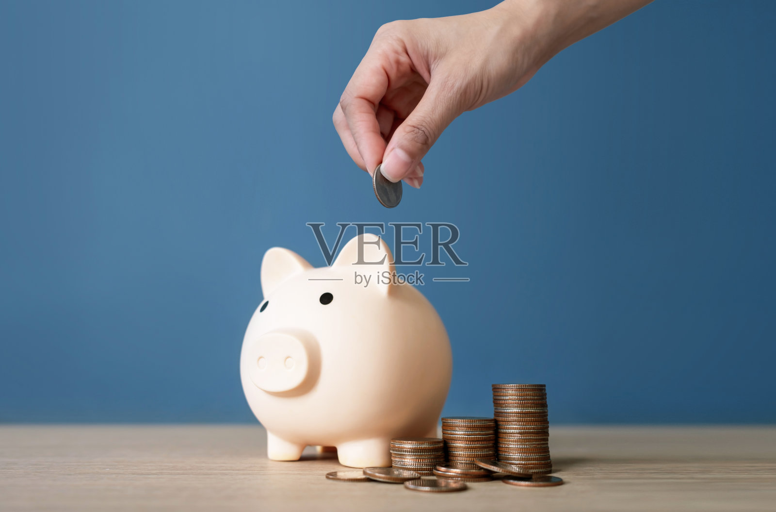女人手将硬币放入木桌上的储蓄罐，背景为蓝色。储蓄和投资概念。照片摄影图片