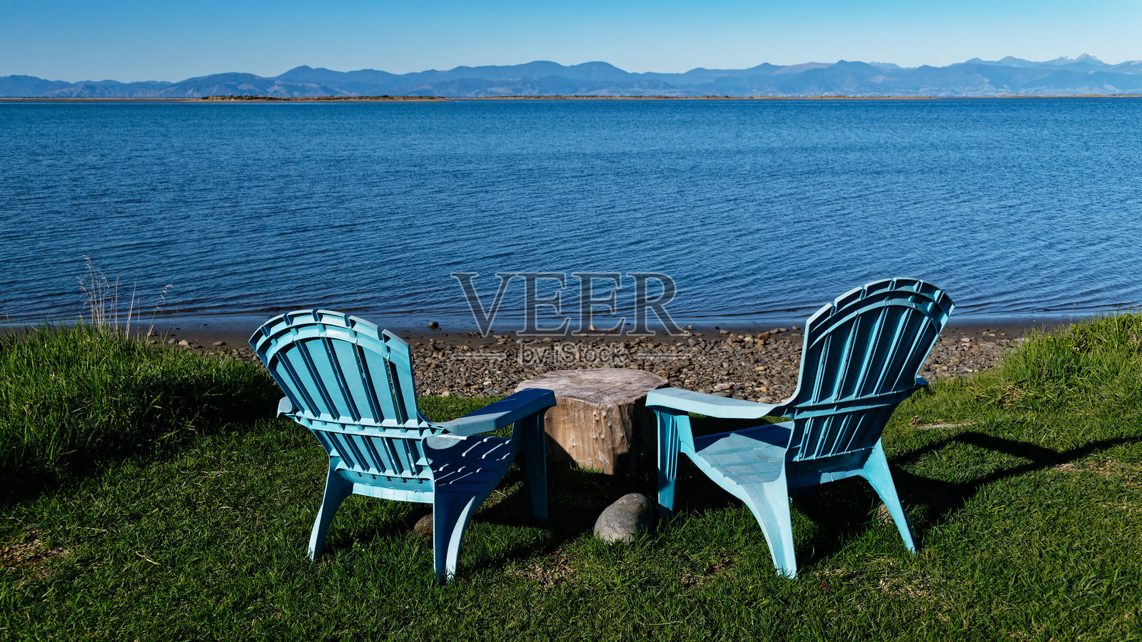 海边草地上放着两把塑料椅子，它们静静地等待着人们坐下，中间摆放着一张由原木制成的乡村风格的桌子。照片摄影图片