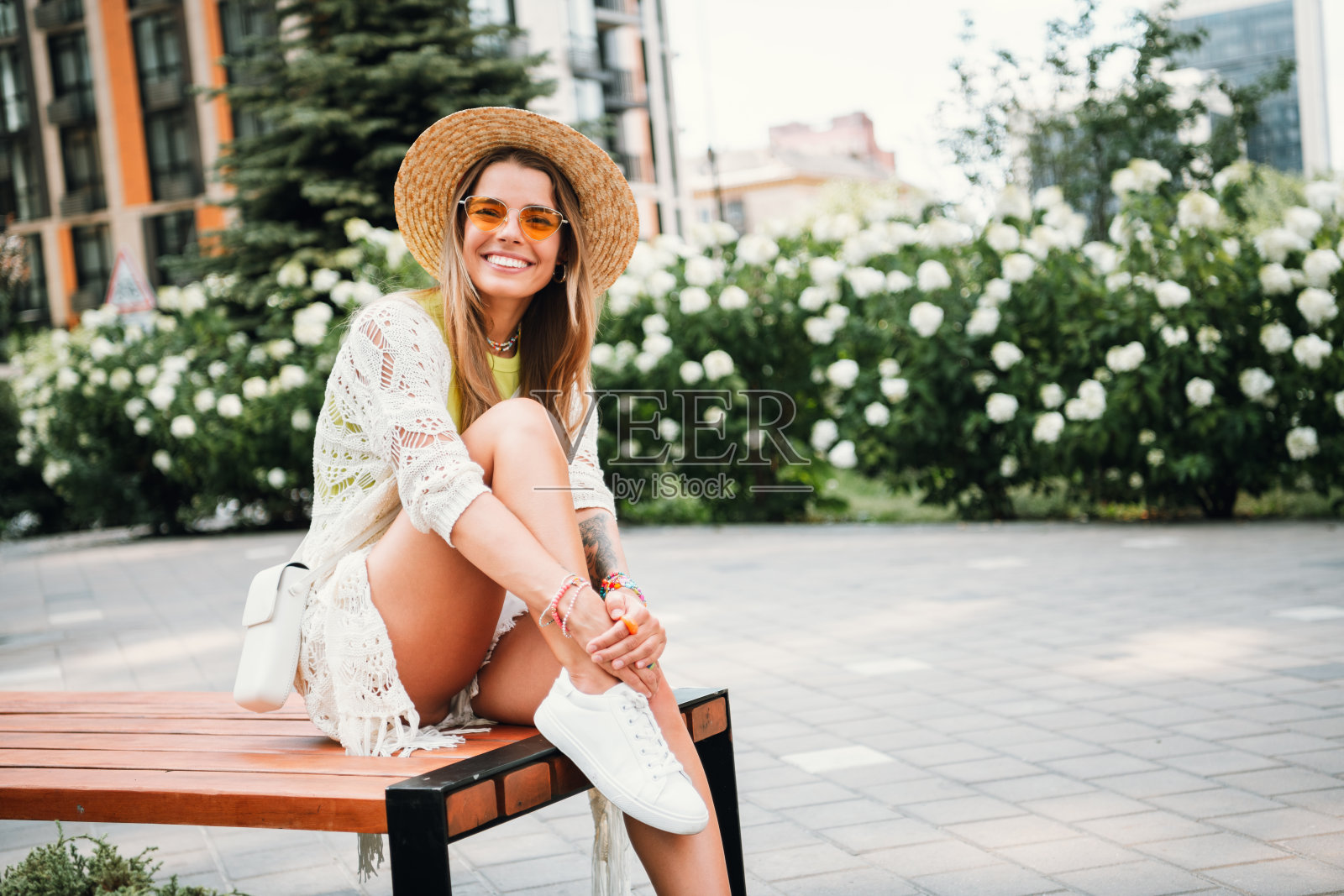 阳光明媚的城市公园里，一位时尚的年轻女子戴着太阳帽和太阳镜，身穿白色蕾丝裙，微笑着坐在长椅上。照片摄影图片