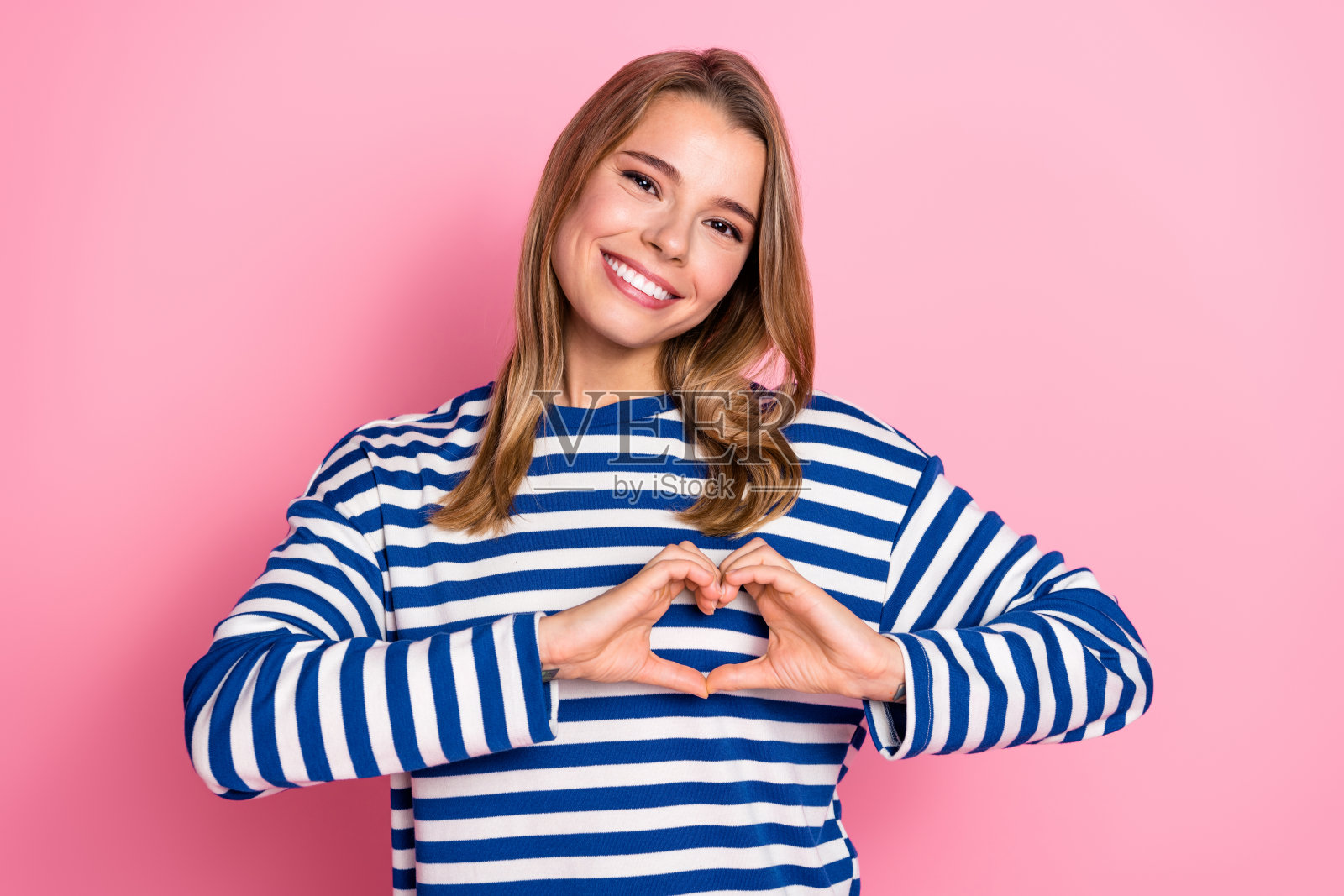 粉色背景上，身穿蓝白条纹毛衣的年轻女子微笑着比出心形手势，展现出幸福与关怀照片摄影图片