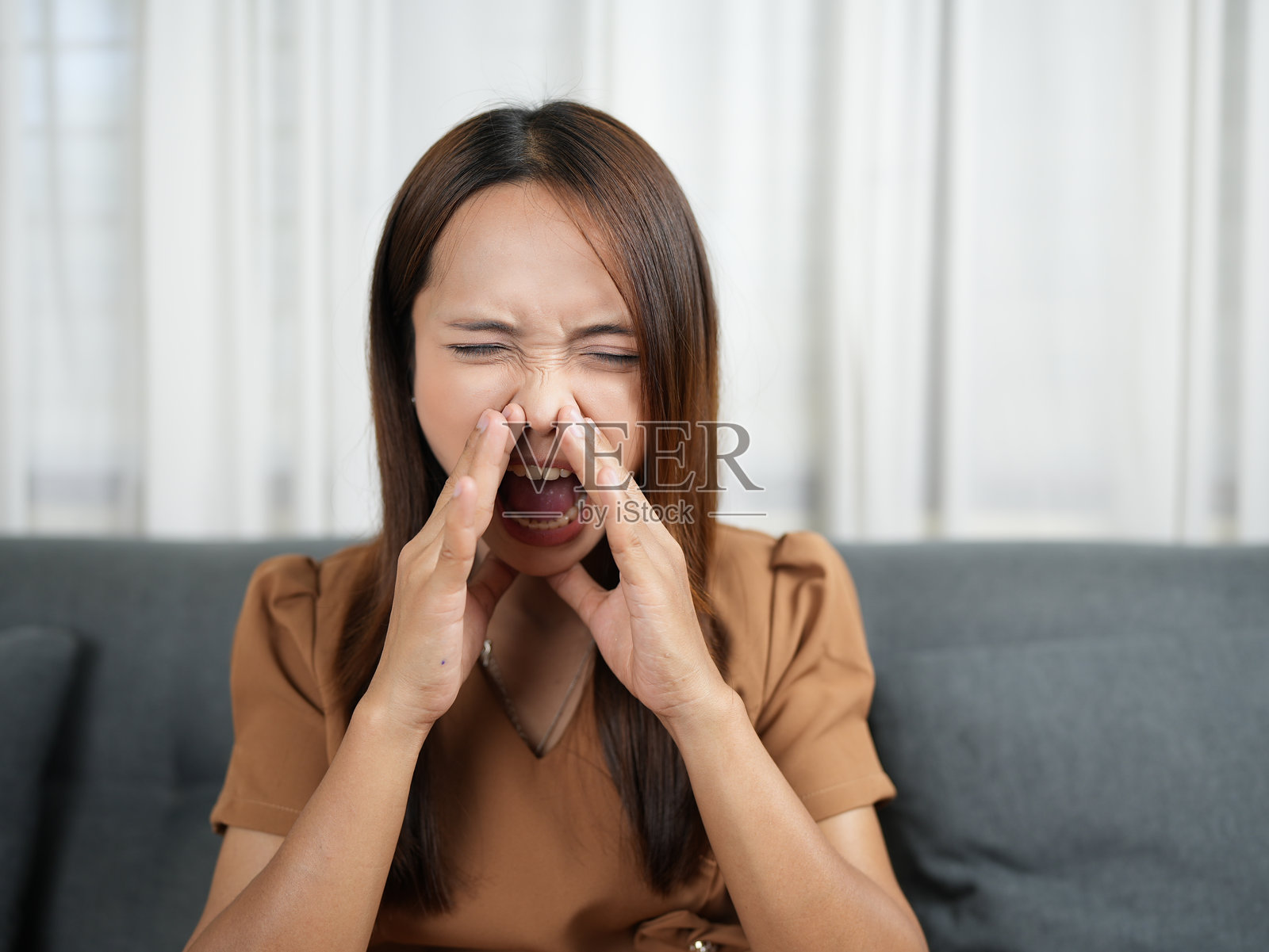 女子坐在沙发上，双手捂脸，闭着眼睛，表达着沮丧，传达出强烈的情绪反应。背景是灯光柔和的舒适室内空间。照片摄影图片