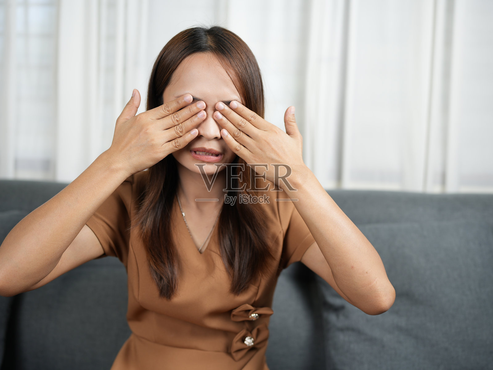 长发女子用手遮住眼睛，表情惊喜或俏皮。她身穿棕色连衣裙，坐在灰色沙发上，周围环绕着柔和的自然光。照片摄影图片