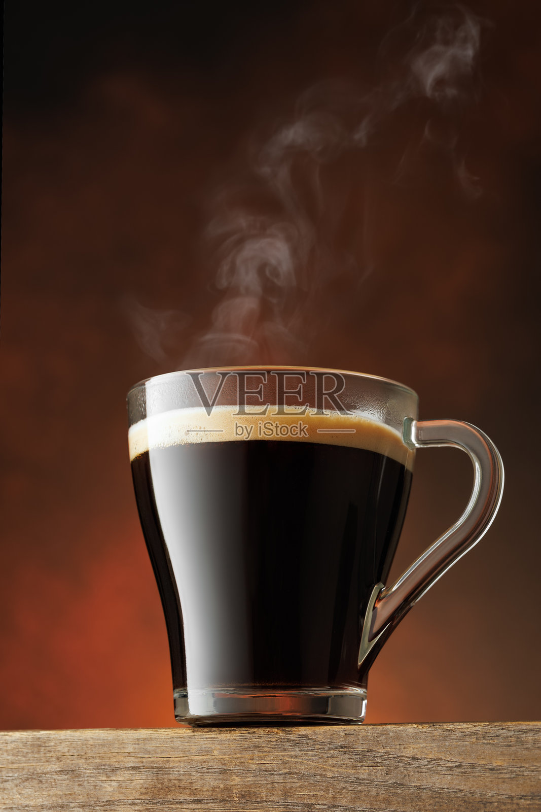 棕色背景下桌子上放着一个透明的热咖啡杯，杯中升腾着热气照片摄影图片