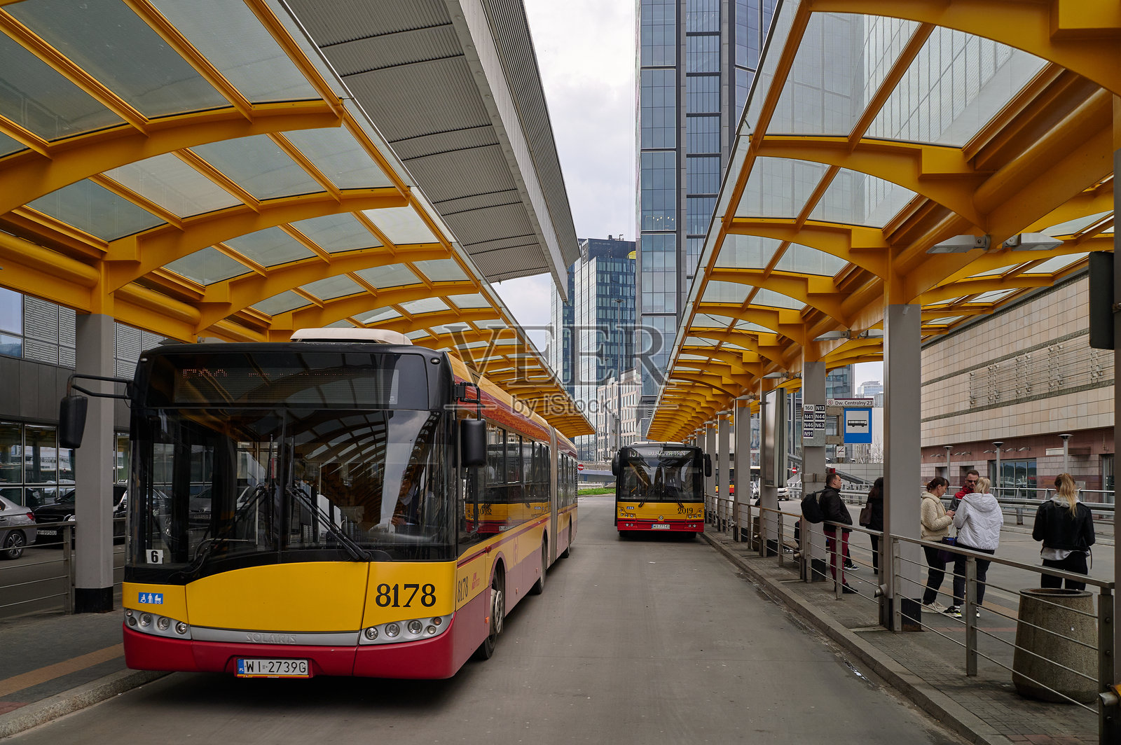 广角镜头下的公交车站，黄色顶棚下停有多辆公交车，乘客们正在等待，展示了现代化的基础设施和市中心的都市景观，白天拍摄，具有建筑构图美感。照片摄影图片