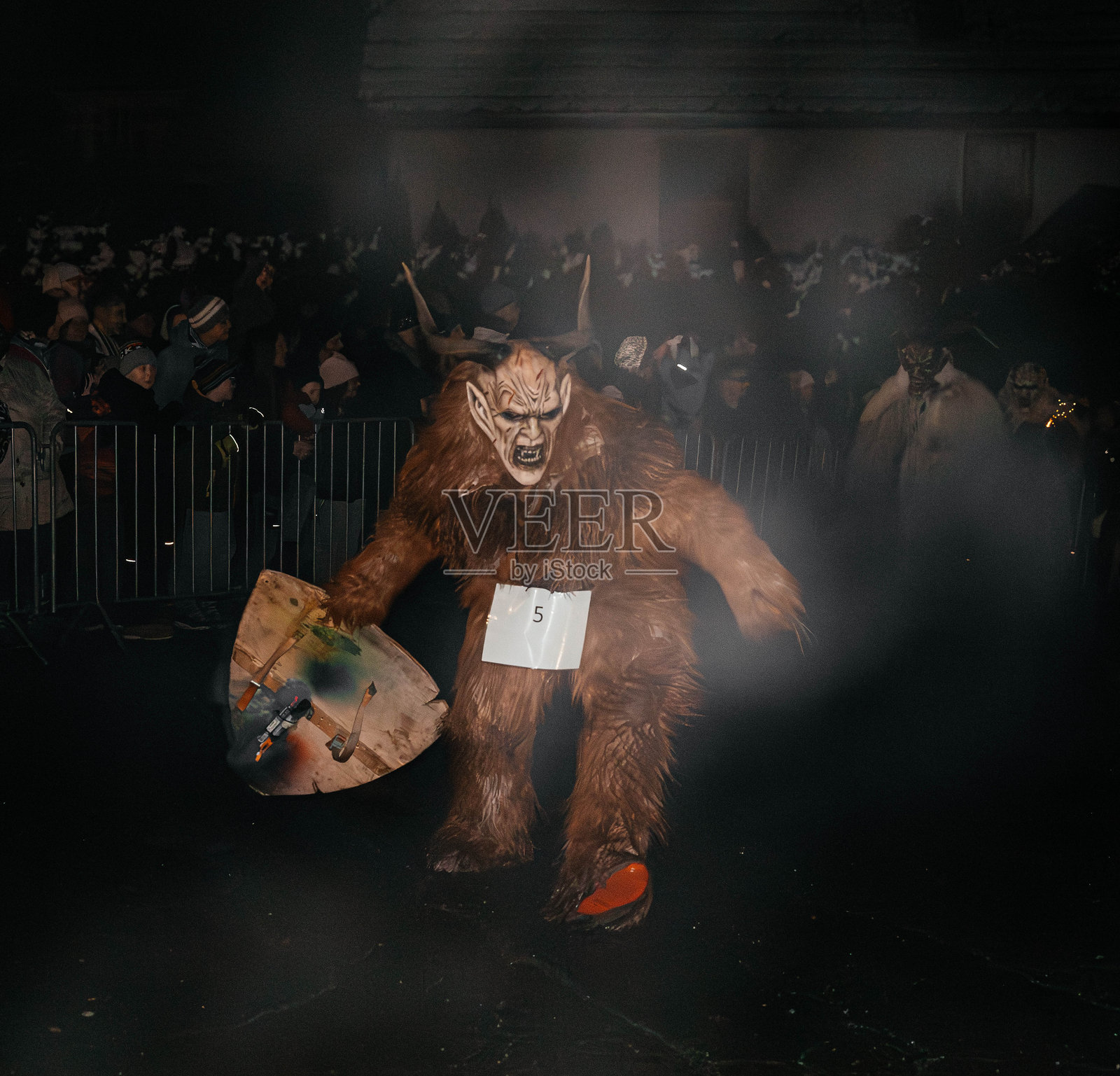 克拉姆佩斯集会诺奇节,节日里,一个打扮成克拉姆佩斯的人身披盾牌,周围是人群,气氛欢腾,夜色朦胧。照片摄影图片