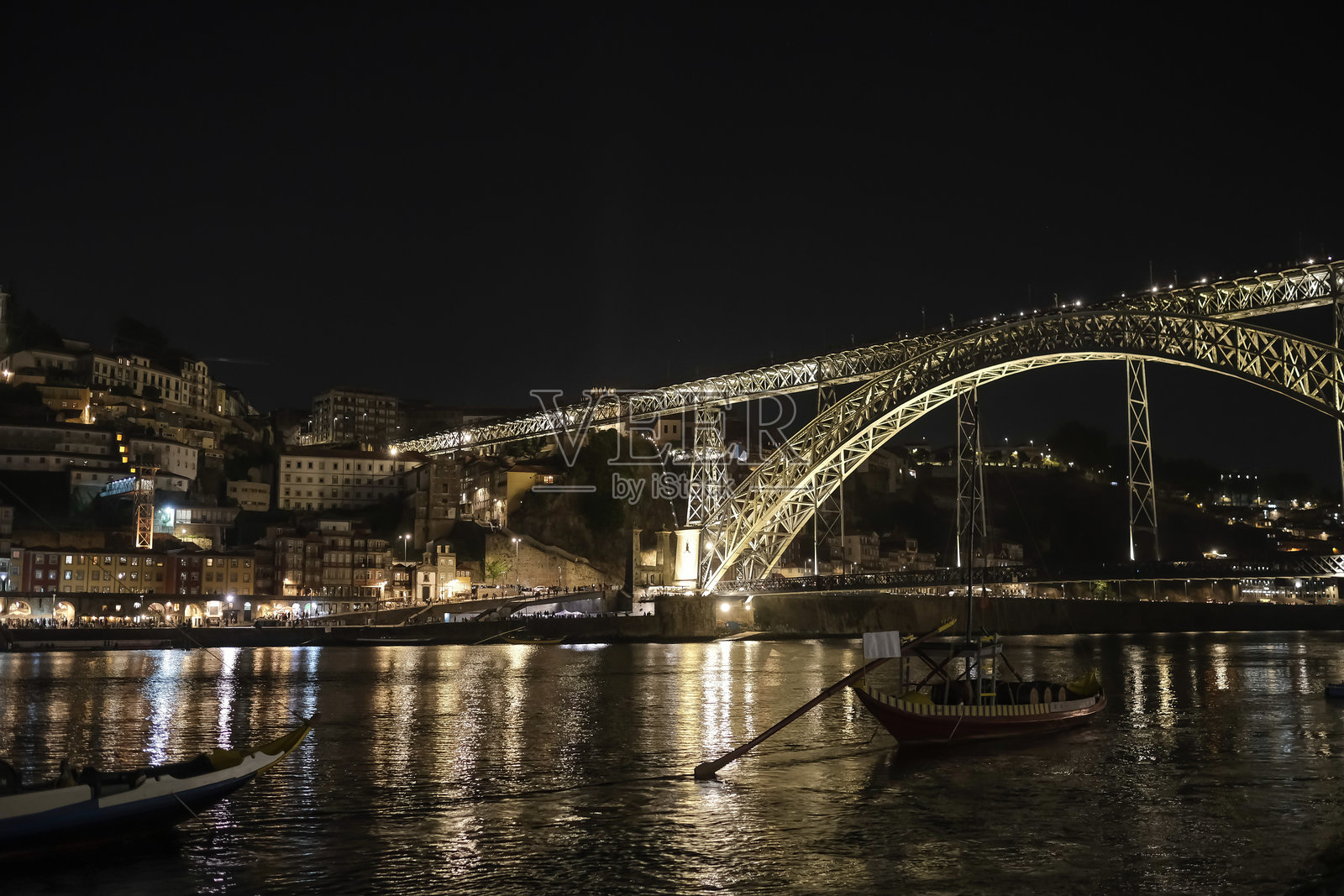 标志性的唐路易斯一世大桥和夜色中的波尔图市景。灯火辉煌的桥梁横跨杜罗河，传统的Rabelo船漂浮在水面上，倒映着金色的灯光。照片摄影图片