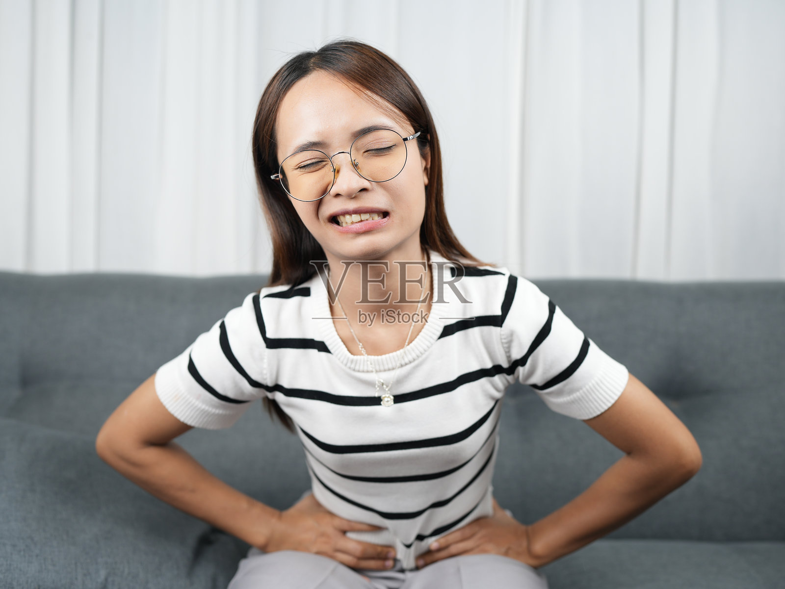 年轻女子坐在沙发上，表情痛苦地捂着肚子，显得很不舒服。她戴着眼镜，穿着条纹衬衫，传递出一种痛苦的感觉。照片摄影图片