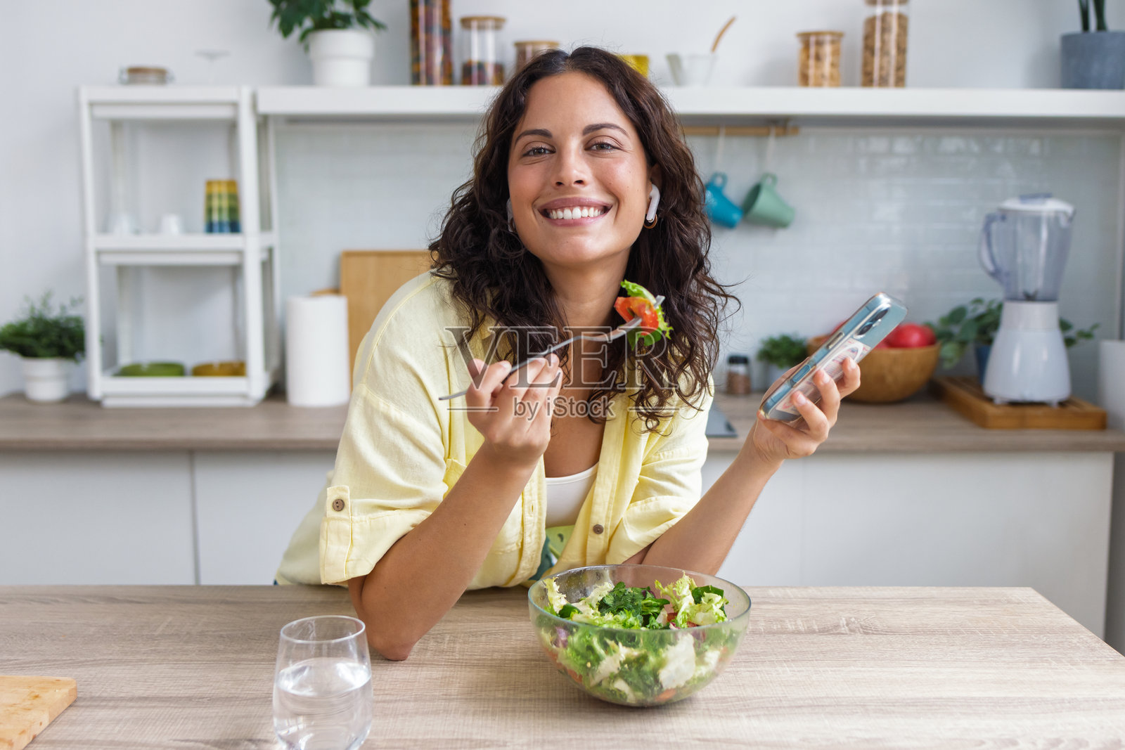 一位美丽的女子在家里的厨房里，一边使用智能手机一边吃着健康的沙拉照片摄影图片