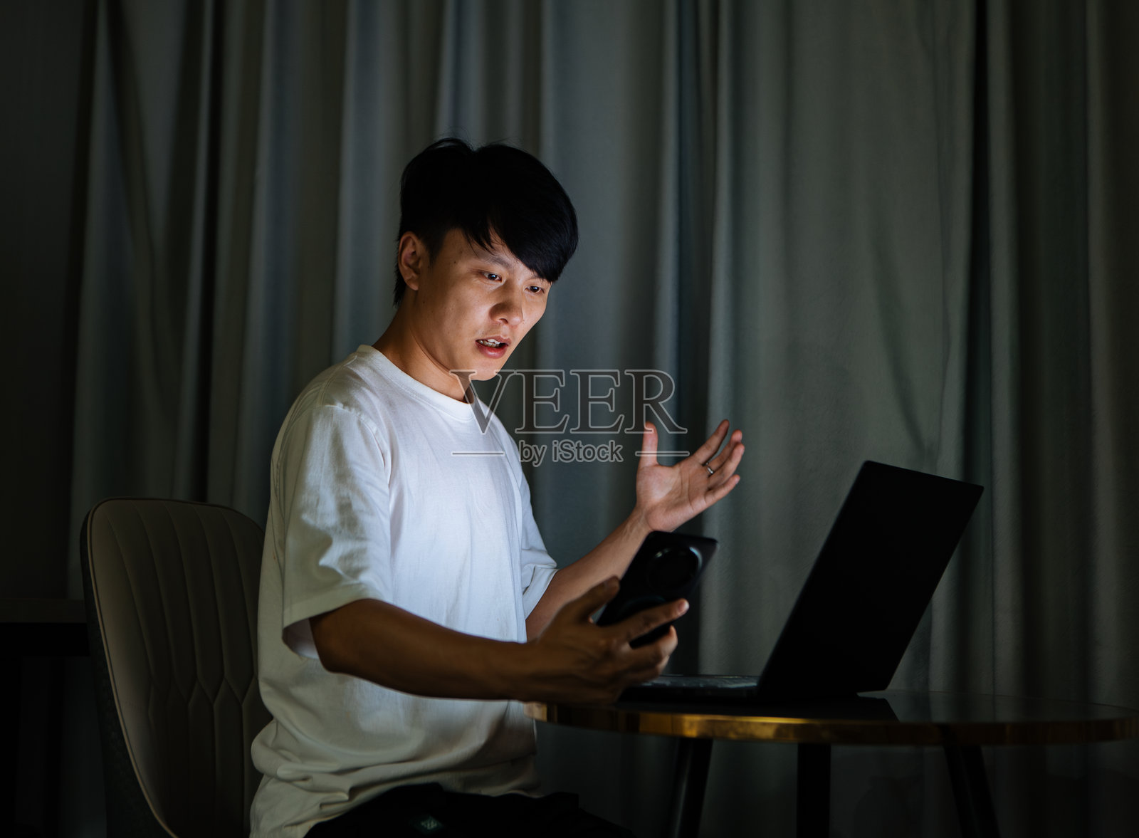 年轻的亚洲男子在家中夜晚使用电脑和智能手机进行视频会议，积极讨论工作，远程协作，并参与现代数字交流和专业的在线会议。照片摄影图片