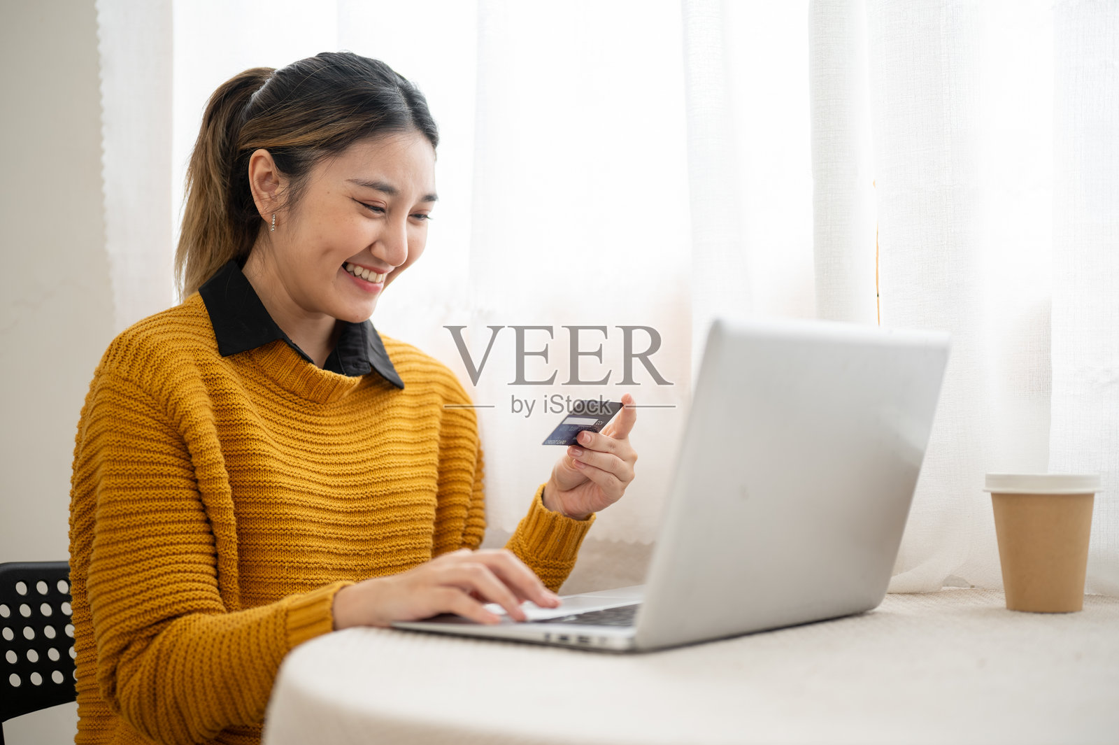 面带微笑的亚洲女性学生或职场人士，手持信用卡，看向桌上的笔记本电脑。照片摄影图片