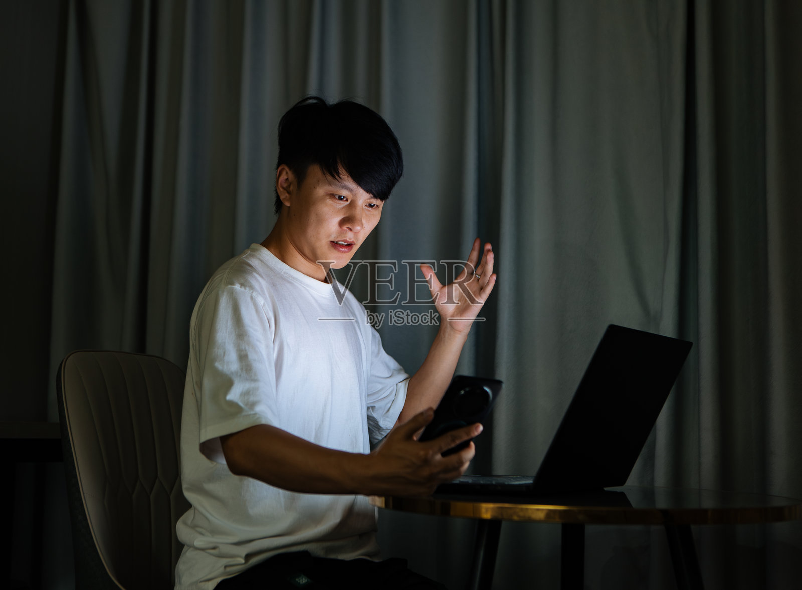 年轻的亚洲男子在家中夜间使用电脑和智能手机进行视频会议，积极讨论工作，远程协作，并进行现代数字通信和专业的在线会议。照片摄影图片