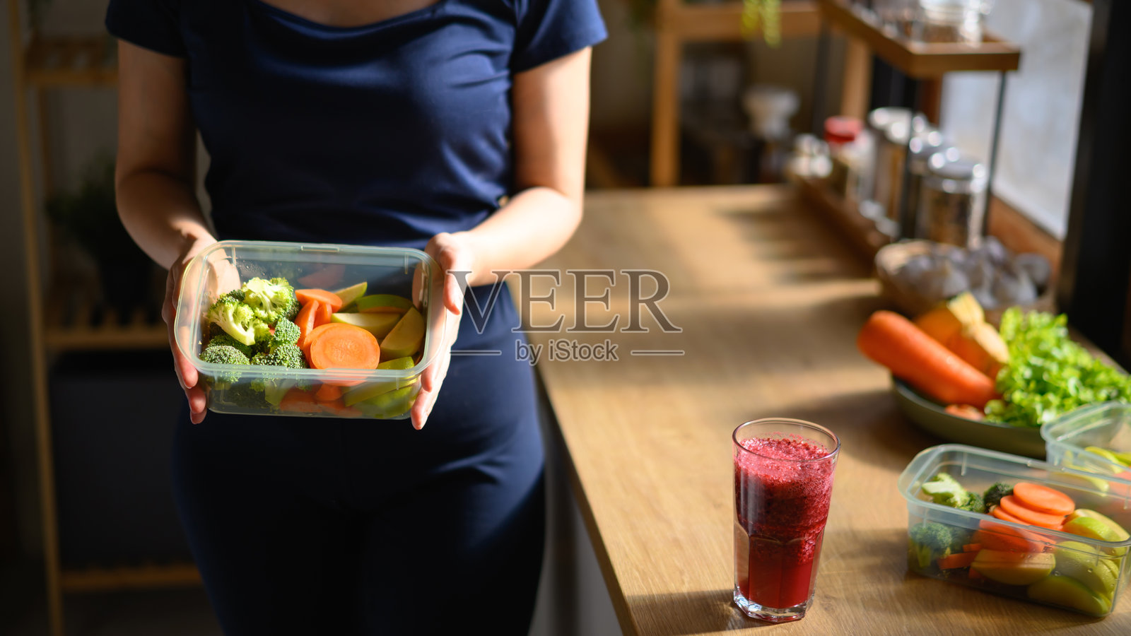 一位女士在家中，桌上放着装有新鲜蔬菜的食物容器和一杯冰沙，象征着健康、用心生活的生活方式照片摄影图片
