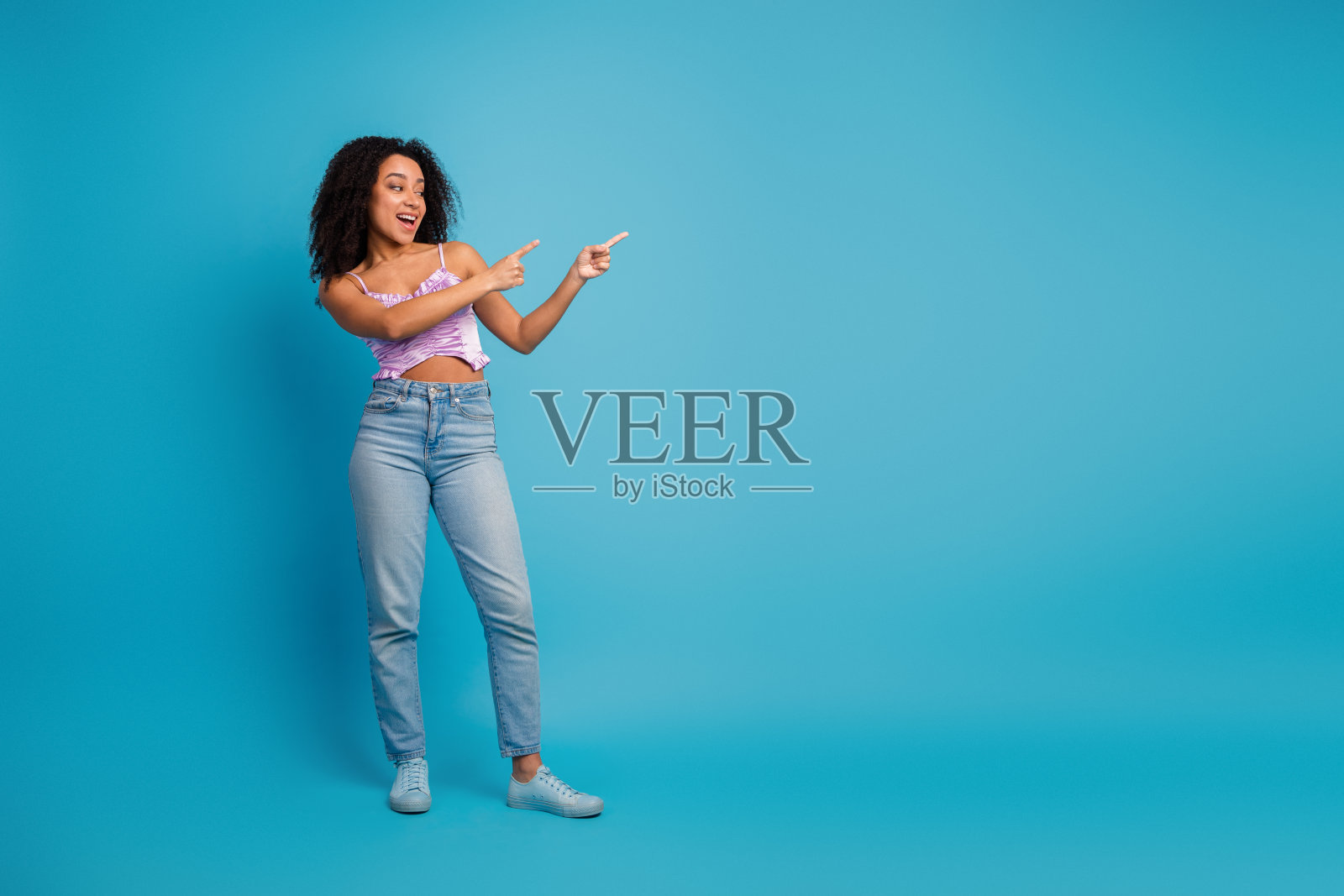 年轻女子卷发，身穿淡紫色上衣和牛仔裤，对蓝色的背景俏皮地伸出手指，展现休闲时尚和快乐的生活方式瞬间照片摄影图片