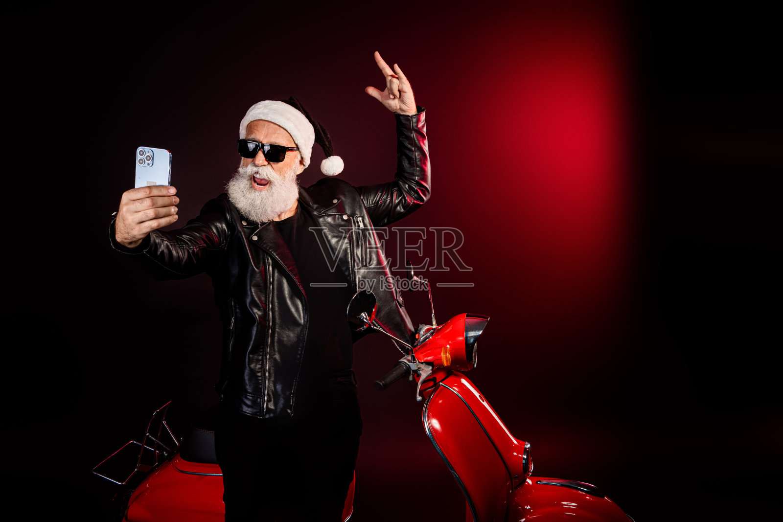 摇滚风的圣诞老人戴着皮夹克和圣诞帽，骑着红色踏板车自拍，在时尚的节日场景中享受圣诞气氛和派对心情照片摄影图片