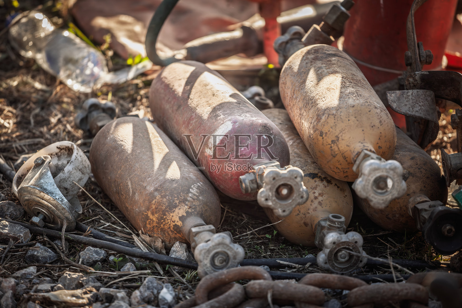 旧的压缩气体钢瓶锈迹斑斑地被丢弃在崎岖的地面上，阀门和软管清晰可见。这些腐蚀的罐子说明了不当处置、工业污染和危险材料风险。照片摄影图片