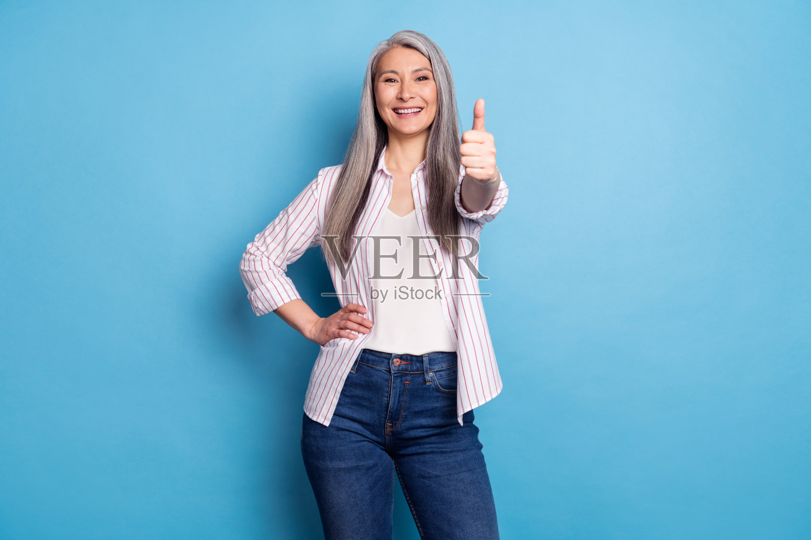 一位迷人的、快乐的、头发花白的女士，竖起大拇指表示信任，她孤立在明亮的蓝色背景上。照片摄影图片