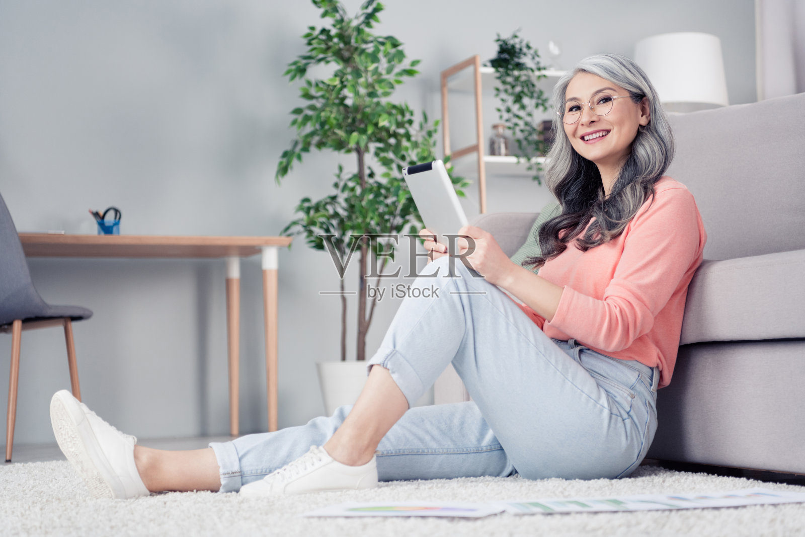 一位漂亮的、和蔼的、快乐的老太太，全身侧面照，她微笑着坐在家里的地毯上，手里拿着平板电脑。照片摄影图片