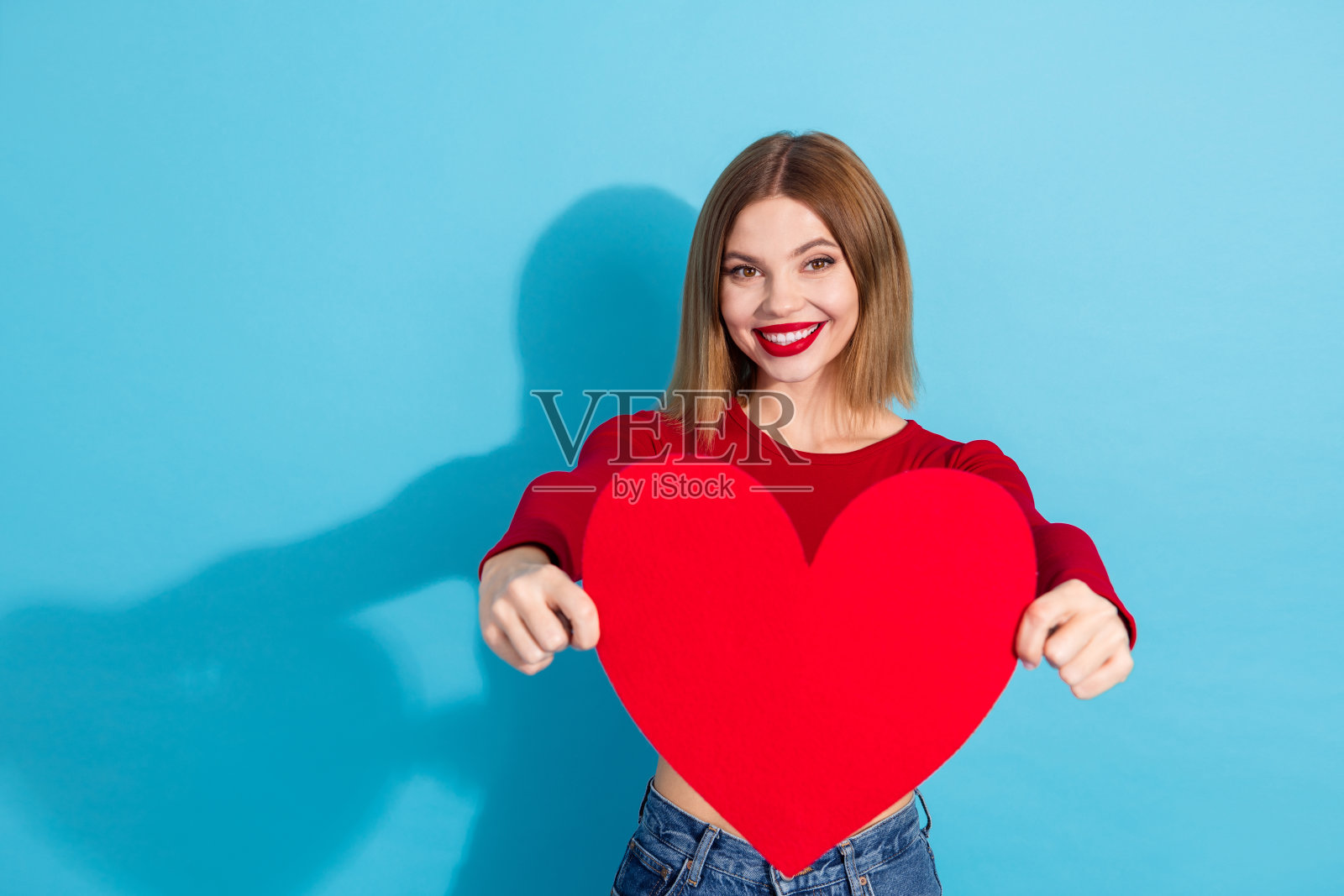 一位身穿红衫的美丽少女，在蓝色背景前抱着一颗巨大的红心，散发着快乐和魅力。照片摄影图片