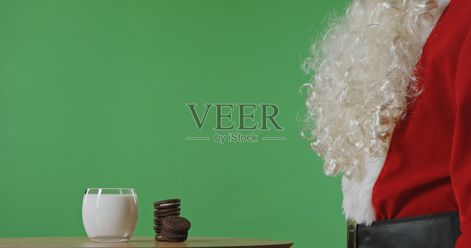 圣诞老人坐在一张桌子旁，桌上放着巧克力饼干和一杯牛奶。照片摄影图片