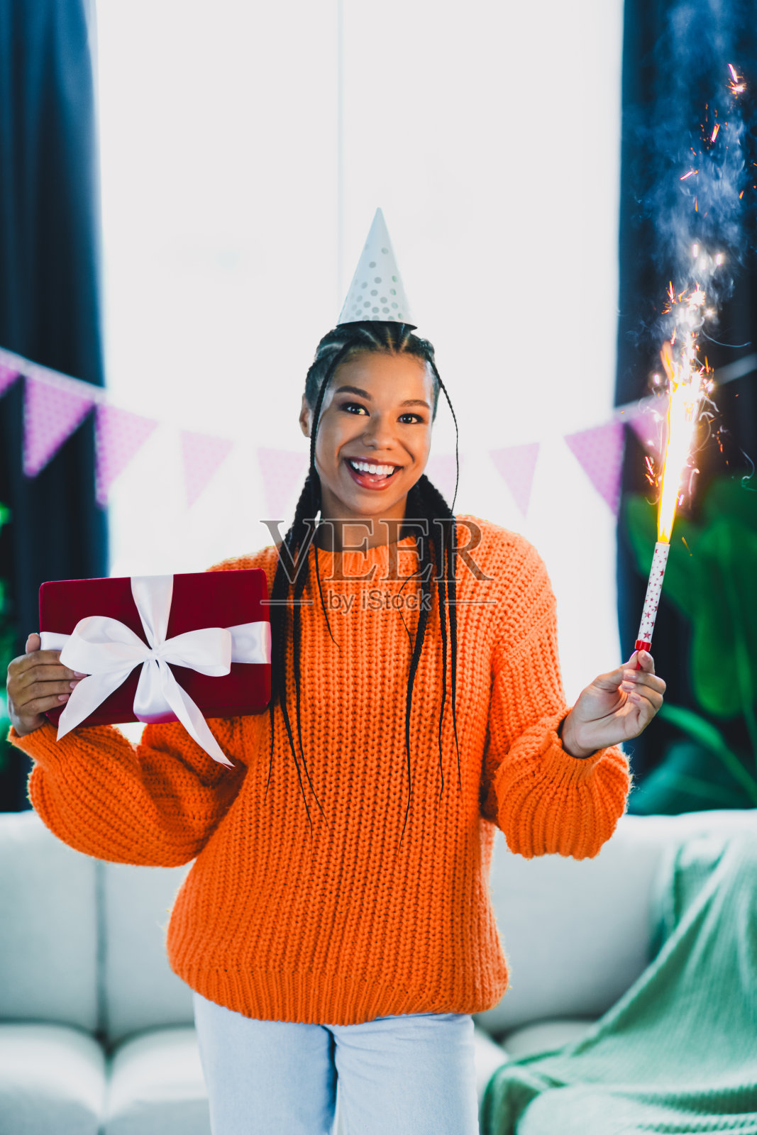 一位身穿橙色毛衣的时尚女士在家中庆祝生日，手持礼物和闪光棒，脸上洋溢着派对的喜悦，朋友们在一旁观看，共同享受温馨的夜晚。照片摄影图片