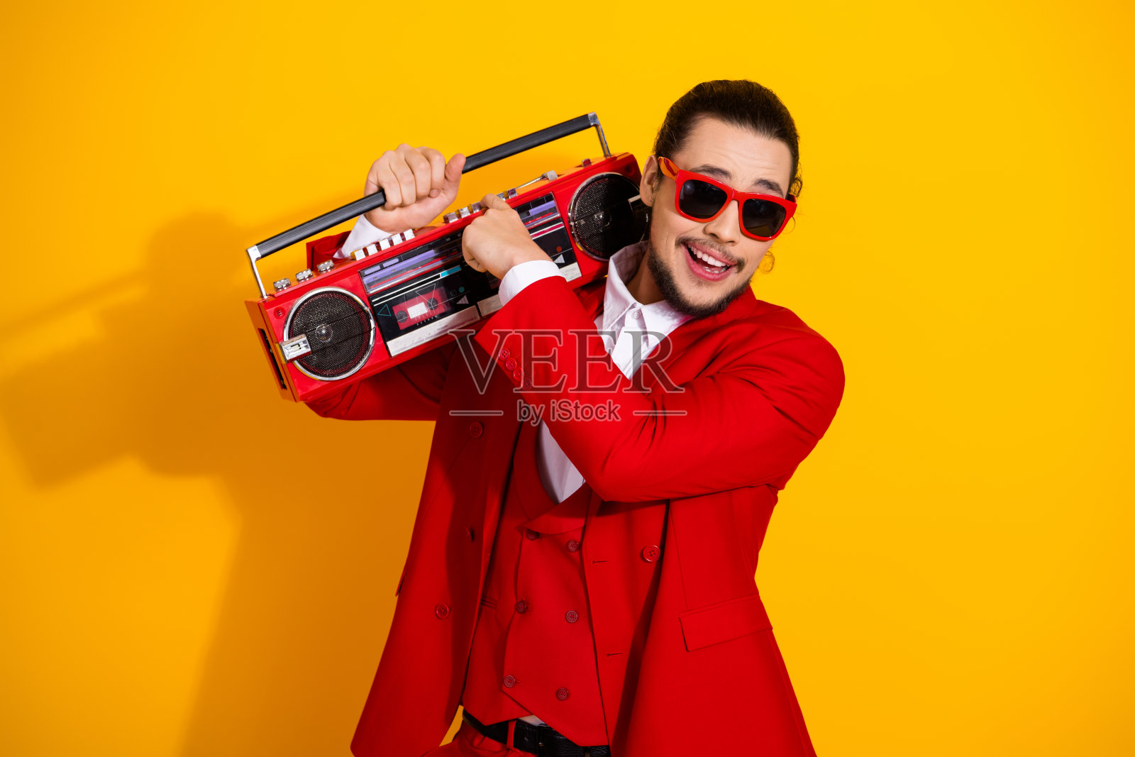 身穿红色西装的滑稽大胖子在明亮的黄色背景下携带复古收音机，用于俏皮的时尚广告照片摄影图片
