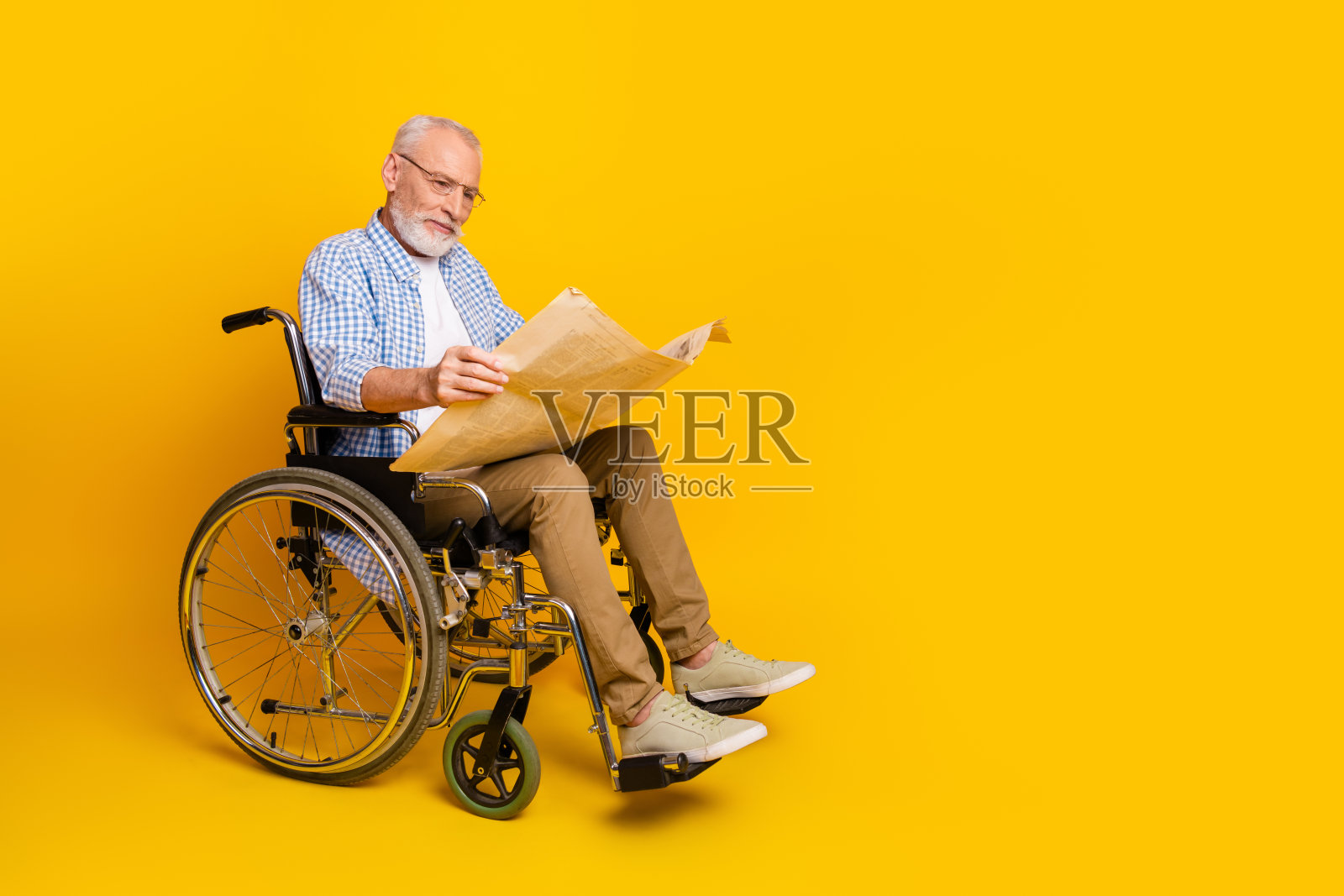 坐在轮椅上的爷爷在明亮的黄色背景下看报纸，展现了康复和支持中的韧性、关爱和日常生活。照片摄影图片