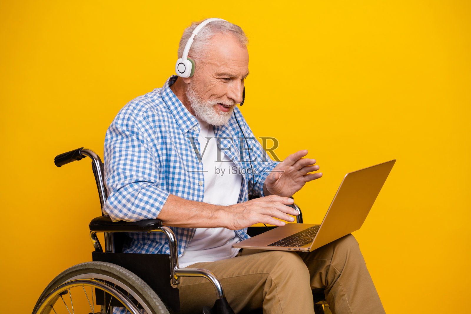 坐轮椅的快乐老人爷爷在阳光明媚的录音棚里，以明亮的黄色为背景，戴着耳机，用笔记本电脑在线学习照片摄影图片