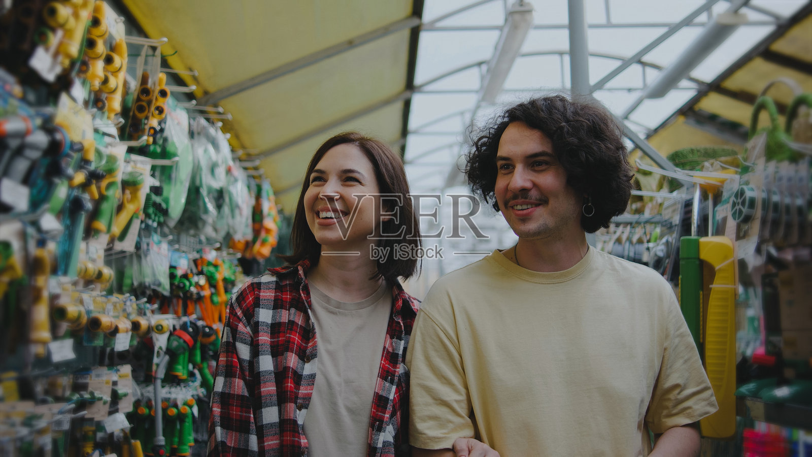 年轻情侣在明媚的日子里，在种满植物和工具的五彩缤纷的温室里，愉快地选购园艺用品。照片摄影图片