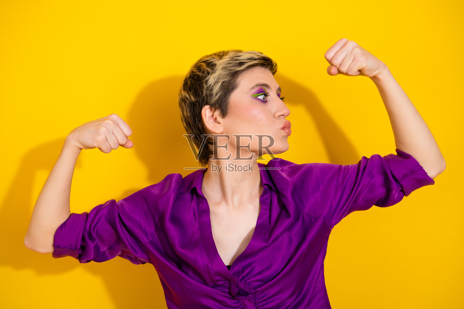 穿着紫色衬衫的自信年轻女子在明亮的黄色背景前展示手臂，展现了迷人的风格和健身活力照片摄影图片