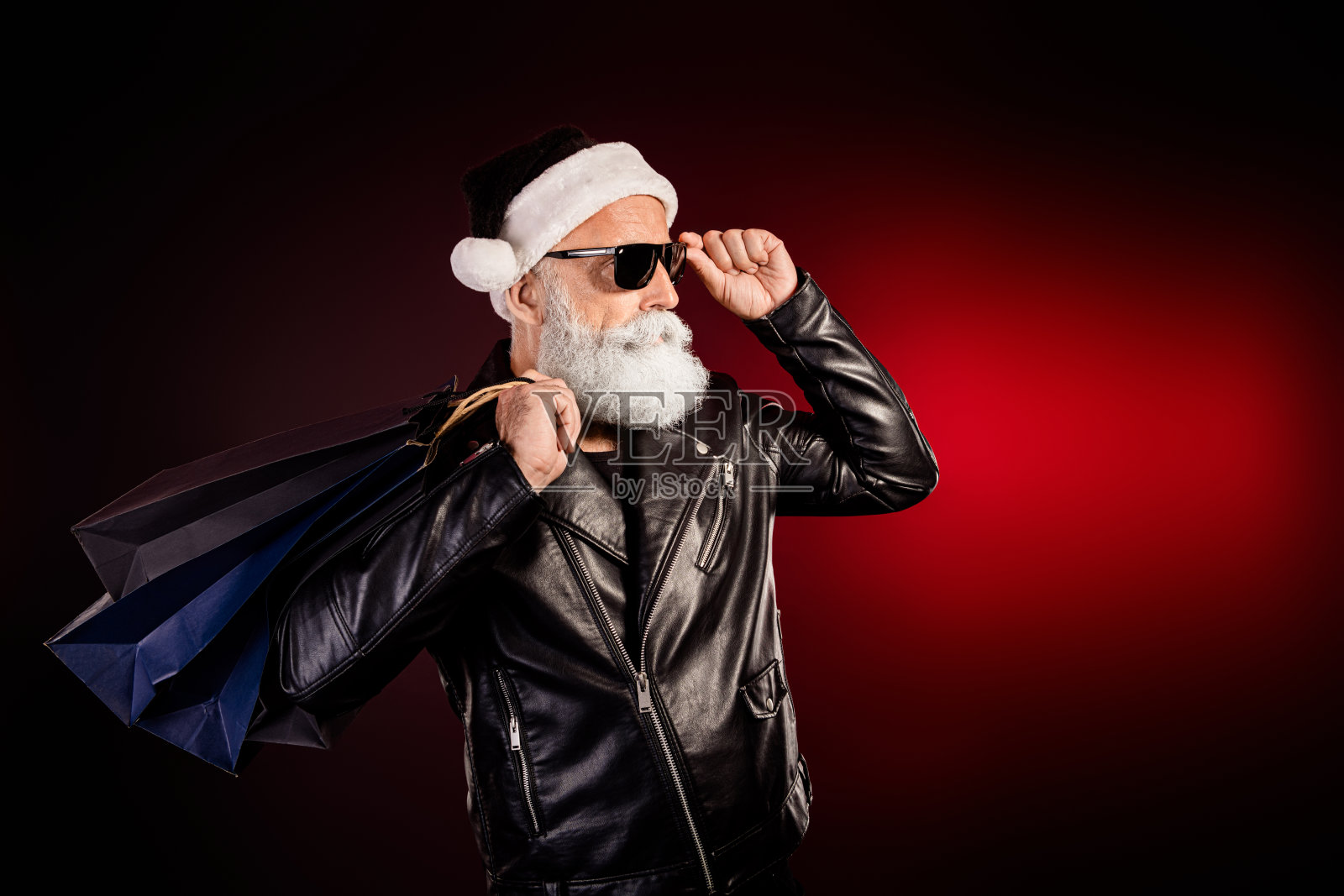 身穿皮夹克、戴着墨镜的摇滚风圣诞老人，提着购物袋，在红灯下摆姿势，为节日圣诞节拍摄的素材照片照片摄影图片