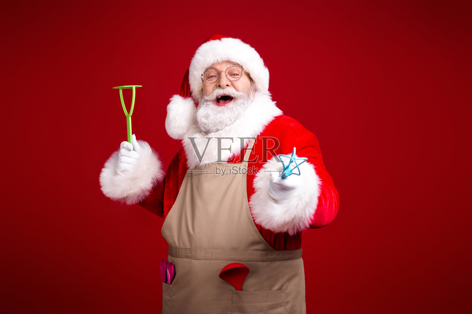圣诞老人身穿米色围裙，手持绿色工具和星形魔杖，在节日气氛浓厚的场景中，以醒目的红色为背景，传递着喜悦。照片摄影图片