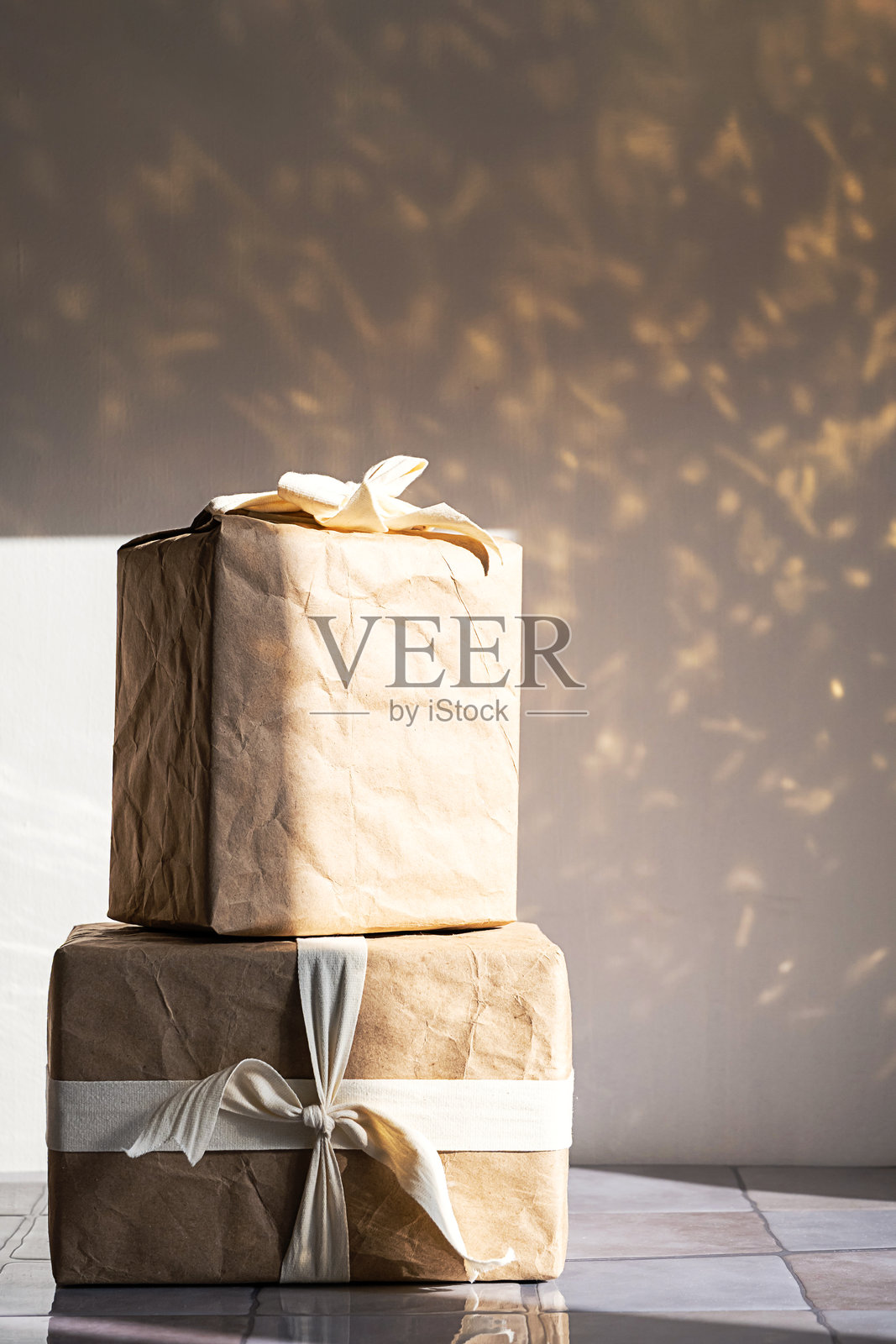 堆叠的礼品盒，用棕色牛皮纸包裹，系着白色丝带，放置在有斑驳光影的反射表面上。照片摄影图片