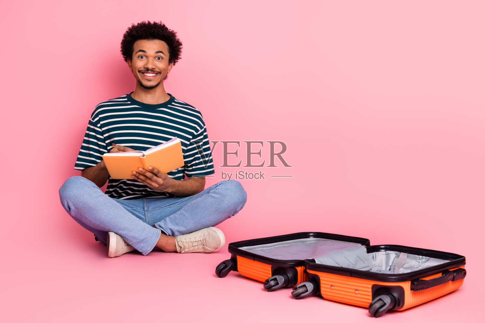 一个年轻的混血男子盘腿坐着看书，旁边放着一个橙色行李箱，背景为粉色照片摄影图片
