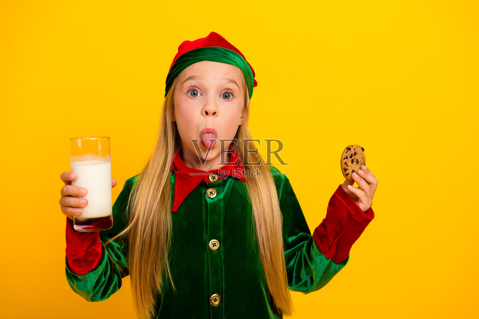 可爱的精灵小女孩扮鬼脸，手拿牛奶和饼干，在节日的黄色背景下庆祝圣诞节照片摄影图片