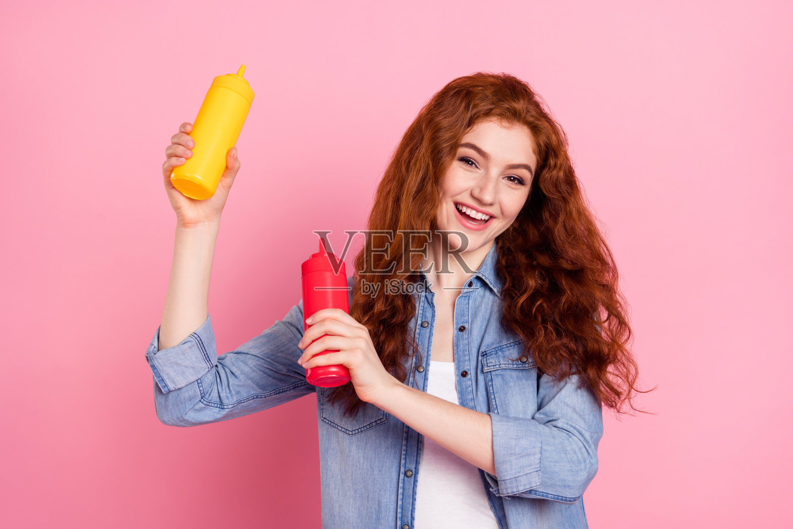 身穿牛仔衬衫的可爱迷人年轻女子，双手举着两瓶酱油，面带微笑，背景为粉色。照片摄影图片