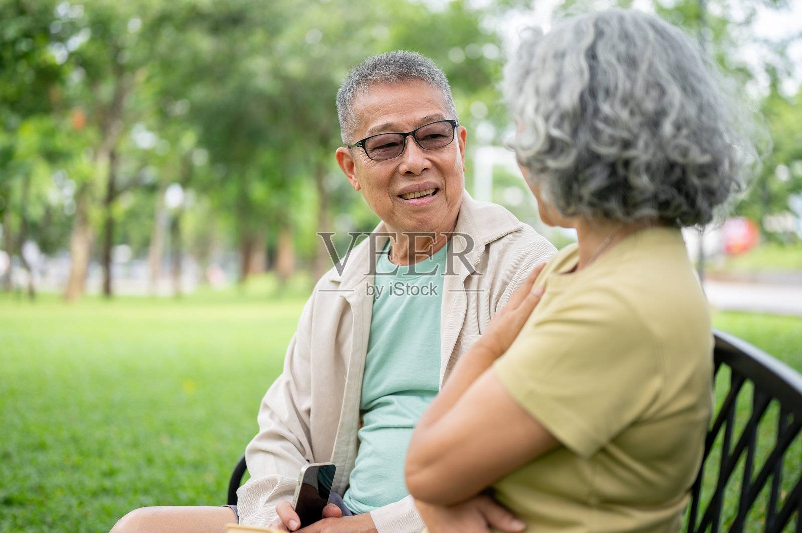 公园长椅上，一位戴眼镜的老人正与一位女士坐在一起，他看着手机，与女士交谈。照片摄影图片